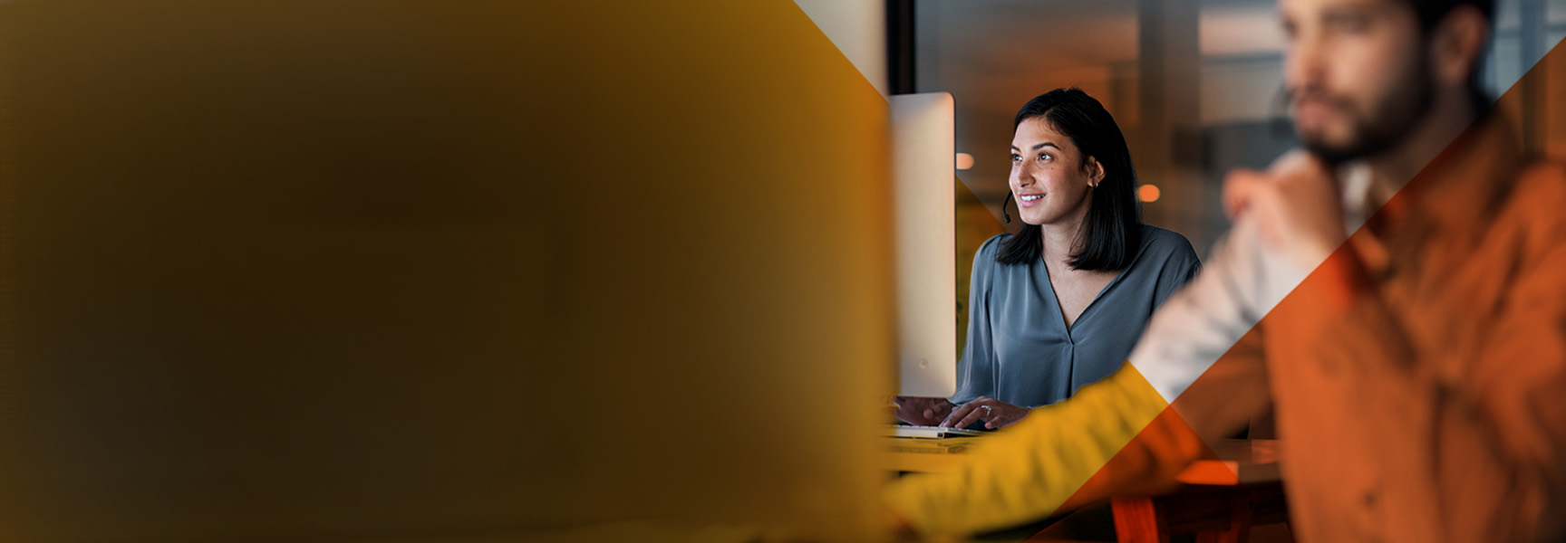 A woman sits at a desk smiling at a computer screen, with a blurred man in the foreground. The scene appears to be in a modern office with warm lighting.