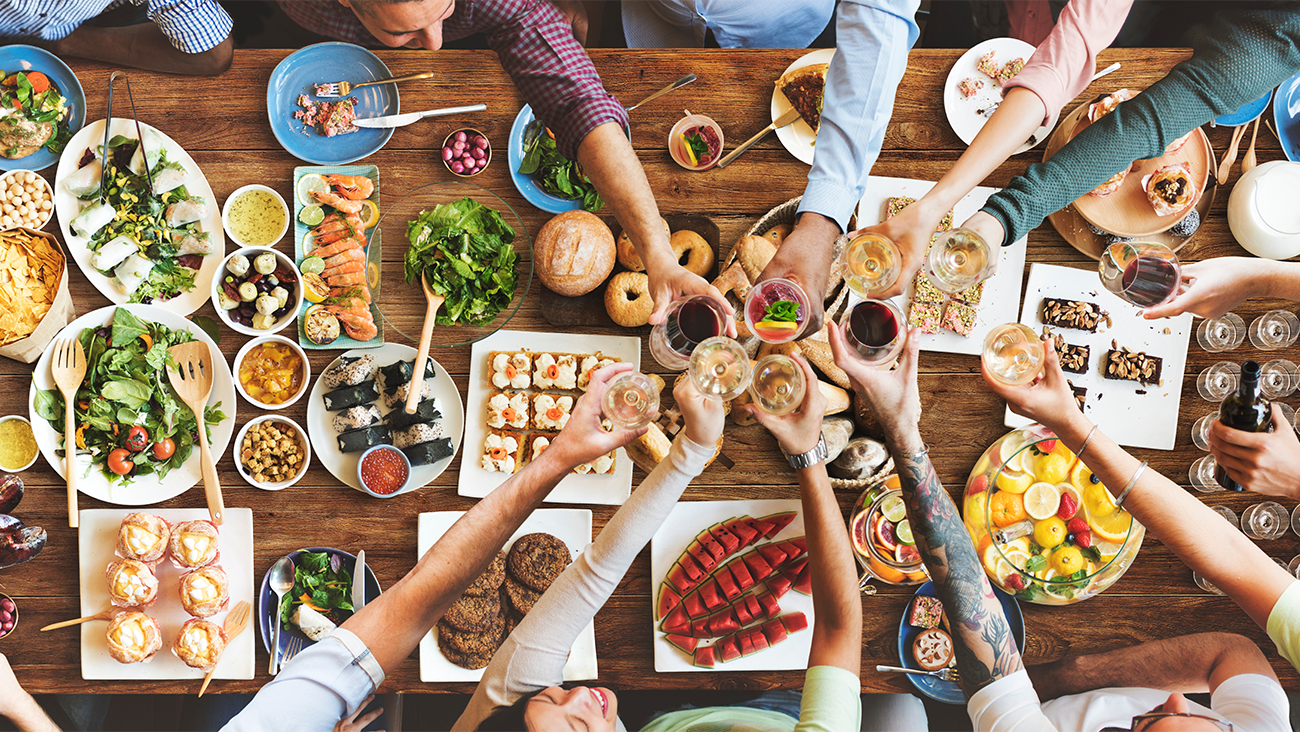A group of people toasting drinks over a table filled with a variety of colorful dishes, salads, sushi, fruit, and appetizers, viewed from above.
