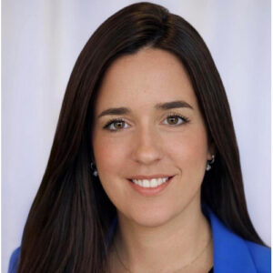 Woman with long dark hair, light skin, and brown eyes, smiling at the camera. She is wearing a blue blazer over a light top and small earrings. The background is plain and softly blurred.